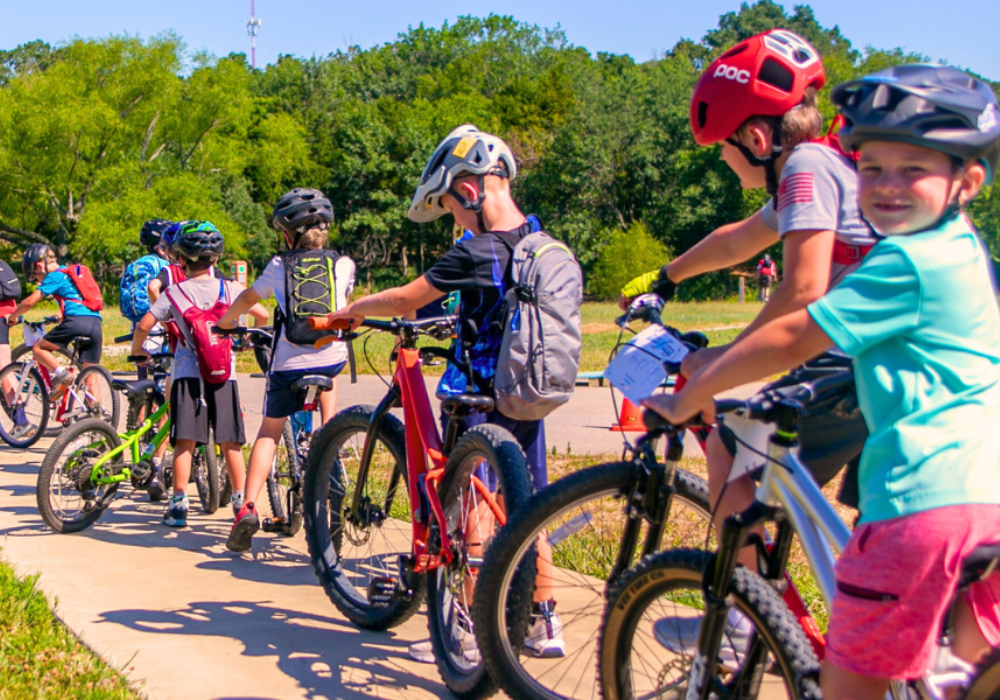 A group of children wearing helmets and backpacks line up with their bicycles on a sunny path, surrounded by green trees and grass—these Pedal Kids are ready for their next adventure.