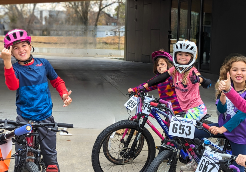 Four Pedal Kids wearing helmets smile and give thumbs up while posing with their bikes, each with a number tag attached. They are outside, near a tunnel, with autumn trees in the background.