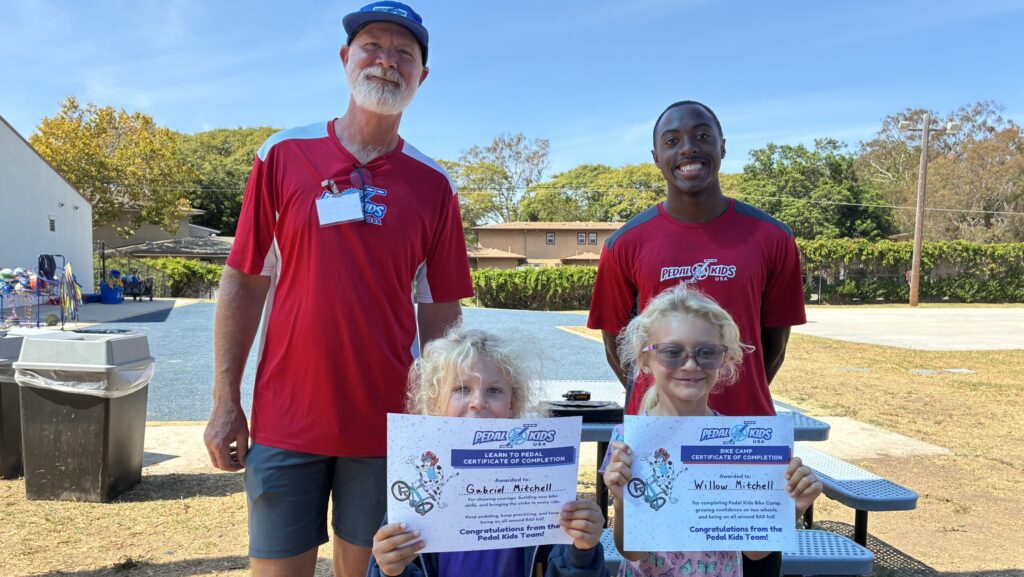 Two adults in red shirts and two children with curly blonde hair—proudly holding their Pedal Kids certificates—stand outdoors on a sunny day. The group is gathered near a bench, with trees and buildings in the background.