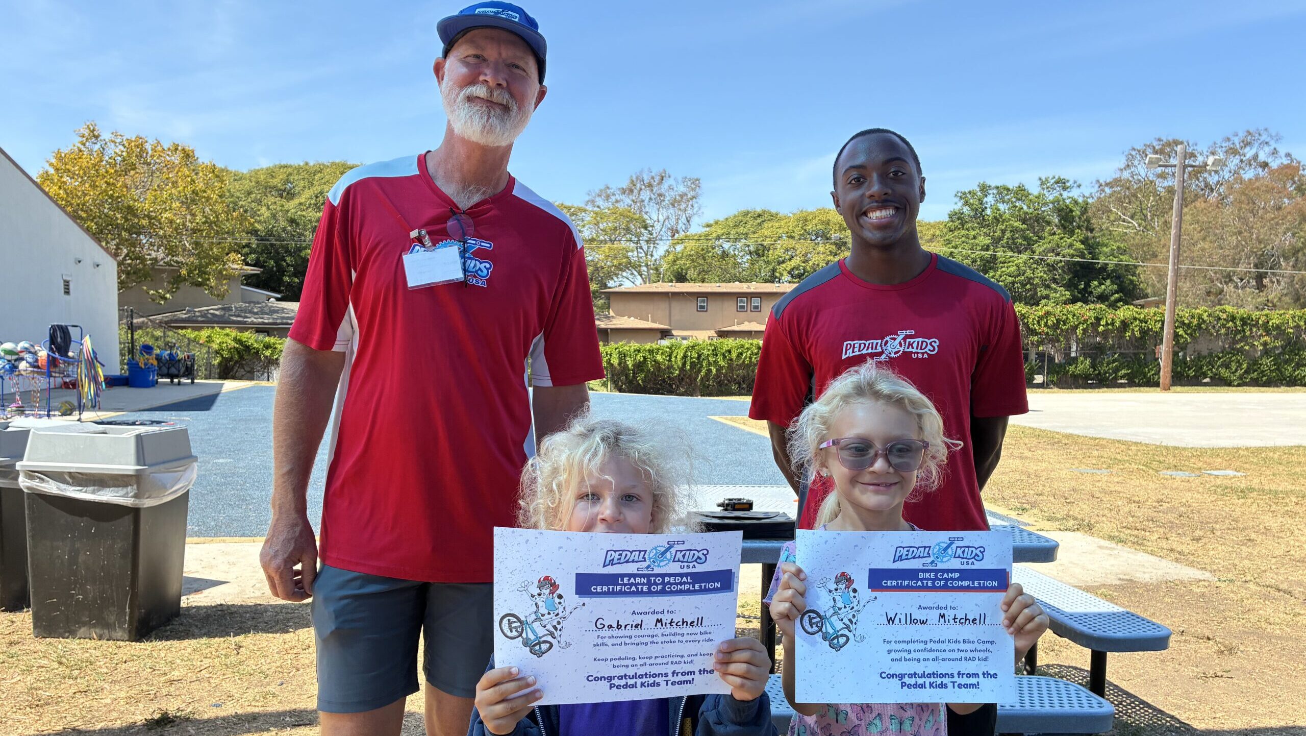 Two adults in red shirts and two children with curly blonde hair—proudly holding their Pedal Kids certificates—stand outdoors on a sunny day. The group is gathered near a bench, with trees and buildings in the background.