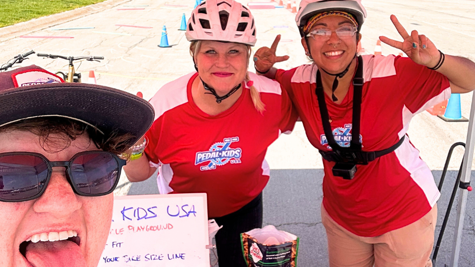 Three people wearing red and white shirts and helmets smile at the camera at an outdoor Pedal Kids bike event. Two show peace signs. A sign reads PEDAL KIDS USA, with cones and bikes visible in the background.