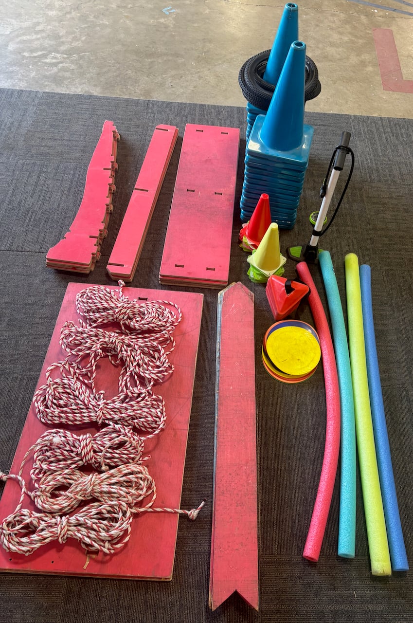 Various gym or playground equipment, including red boards, ropes, blue and yellow cones, an air pump, red and yellow markers, discs, and four colorful pool noodles, arranged neatly on a gray surface.