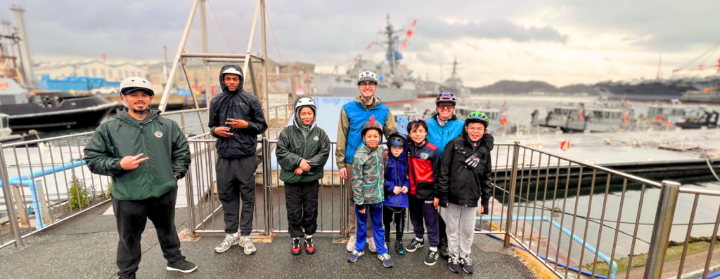 A group of nine people, including adults and children in helmets and jackets, stand smiling on a dock with ships in the background on a cloudy day.