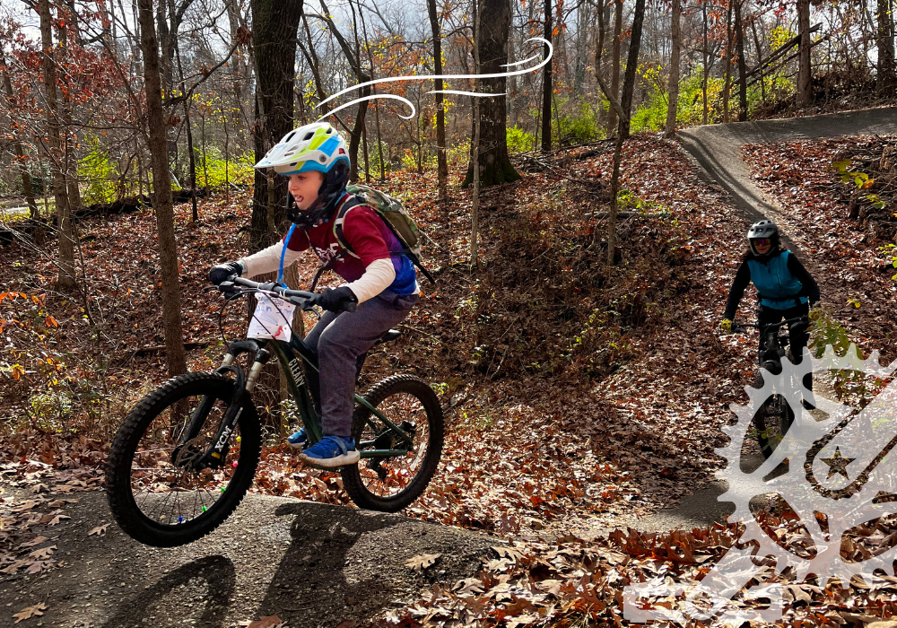A child in a helmet rides a mountain bike on a dirt trail in autumn, with another cyclist following behind.